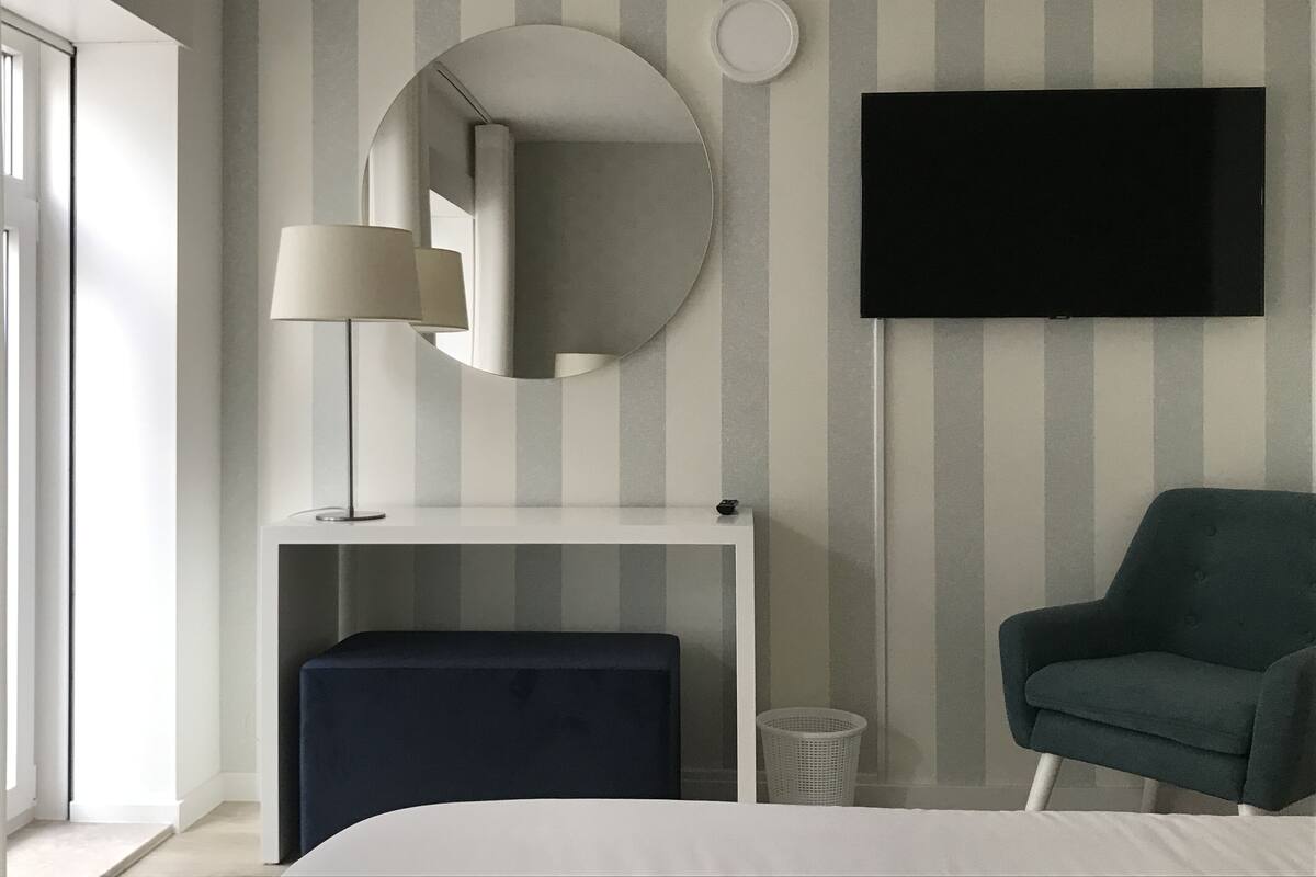 A modern hotel room with a bed, a desk with a lamp, a round mirror, a television, and a dark armchair.