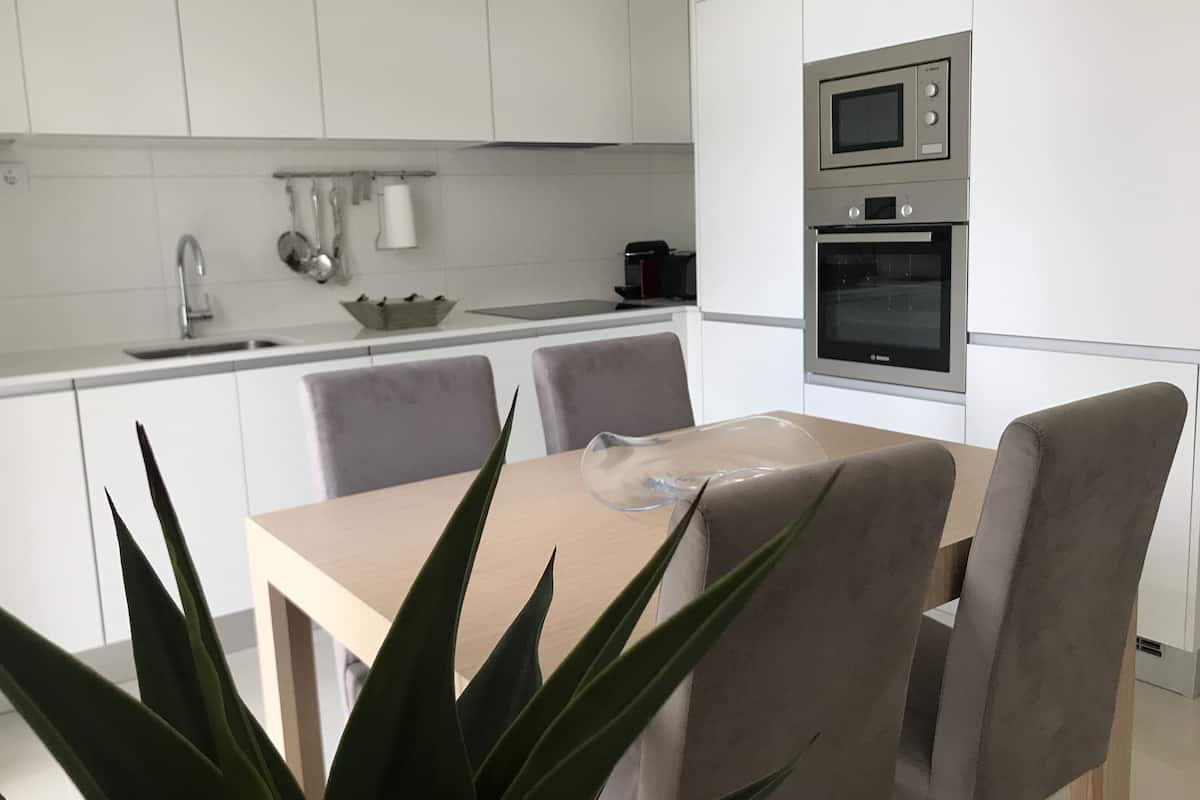 A modern kitchen with white cabinets, a microwave, and an oven. There is a dining table with chairs and a plant in the foreground.