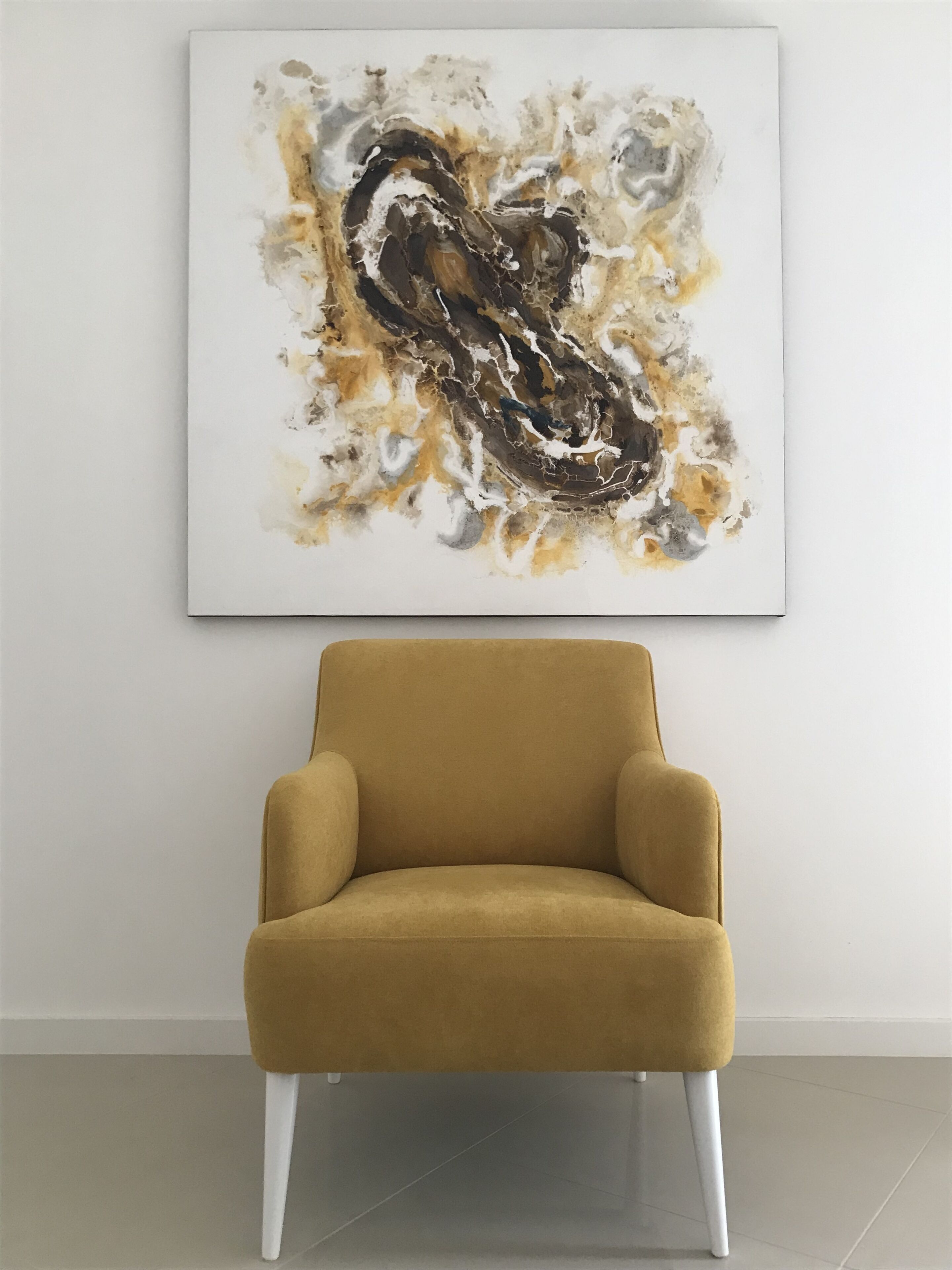 Un fauteuil de couleur moutarde aux pieds blancs, placé devant un mur blanc orné d’un grand tableau abstrait.