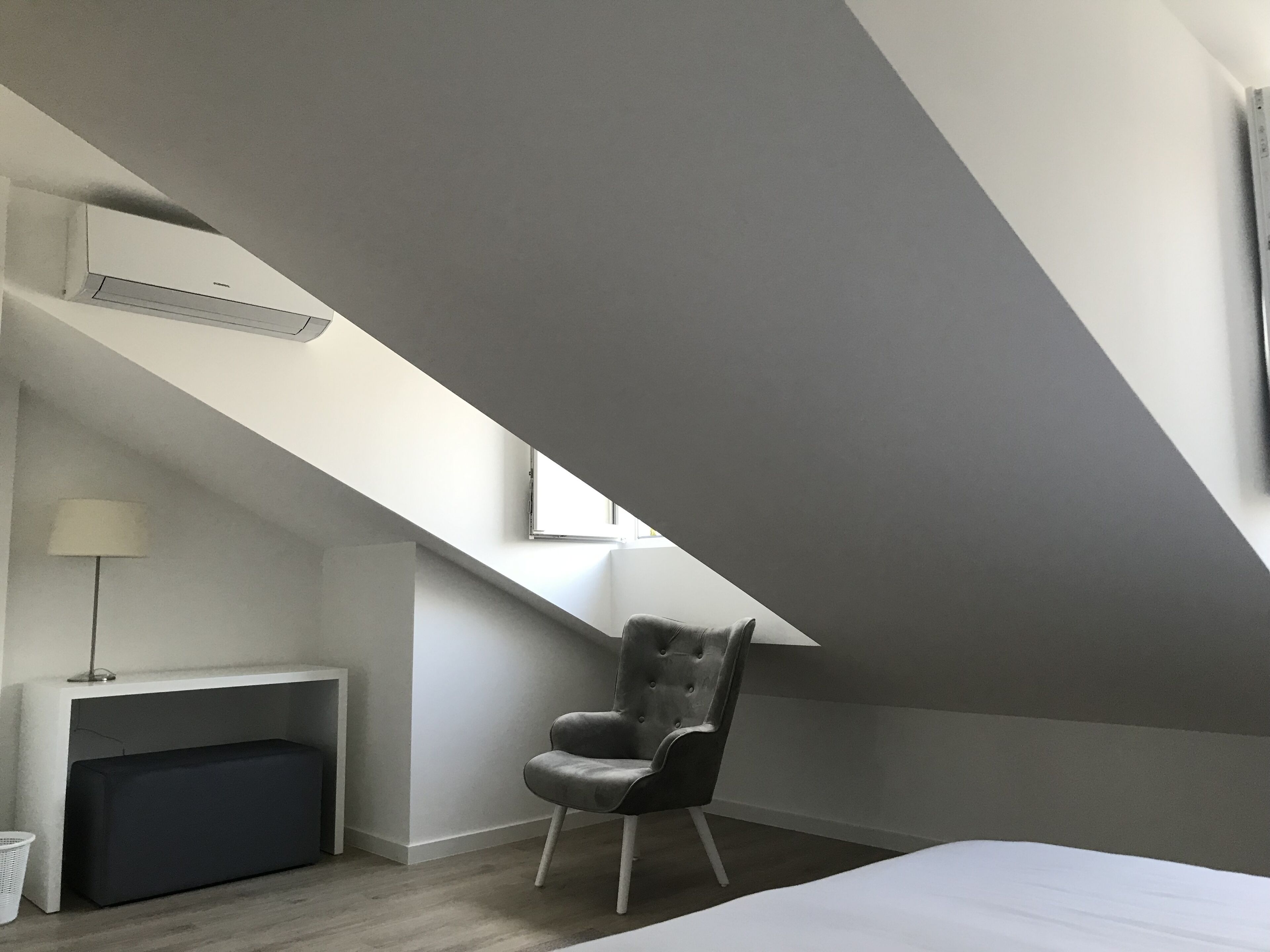 Ein modernes Schlafzimmer mit schräg angelegter Decke, einem grau gepolsterten Sessel, einem Nachttisch mit Lampe und einem großen Bett.