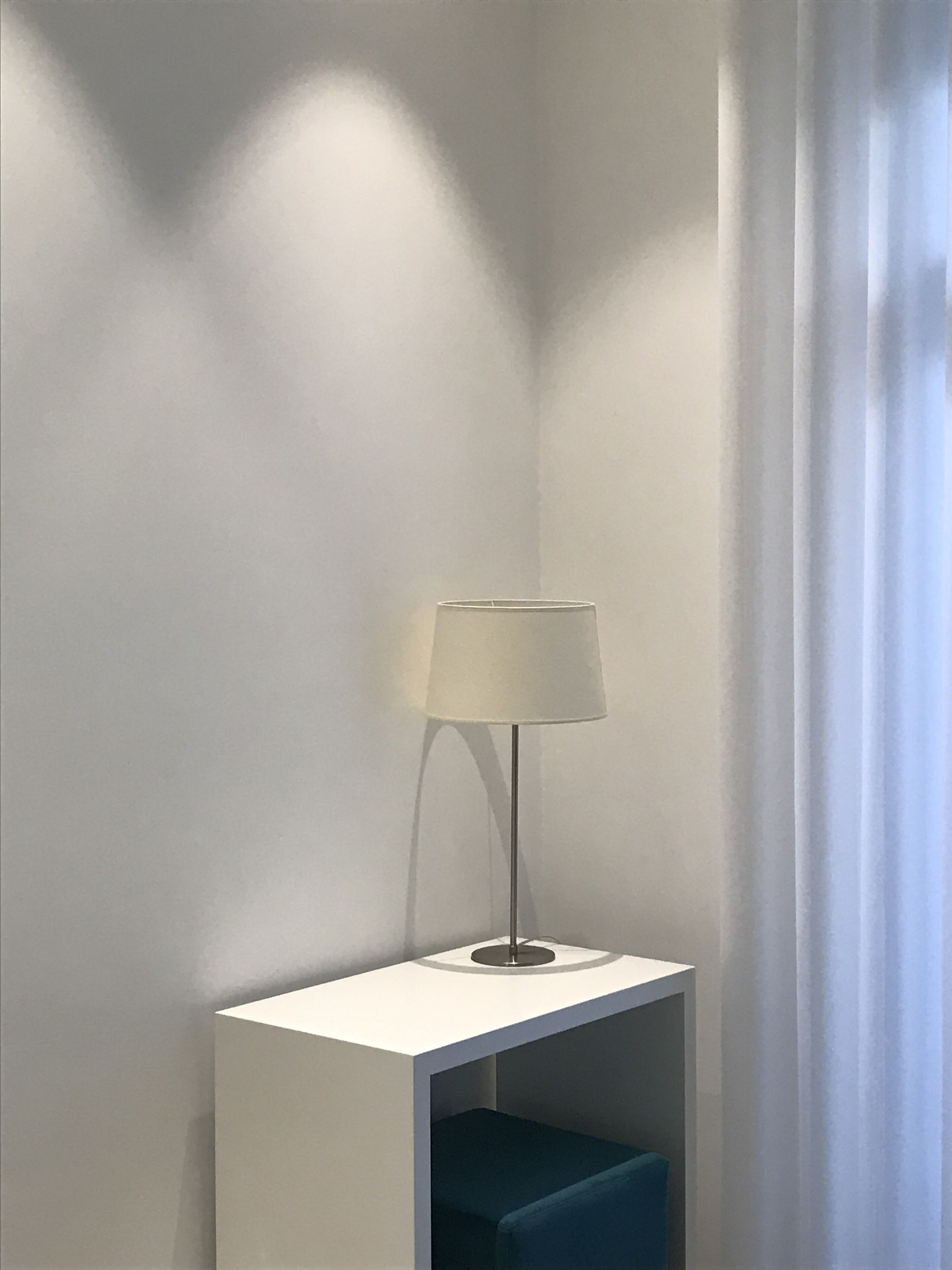 A corner with a white floor lamp on a white pedestal, a blue bench, and a white wall with a shadow cast by the lamp.