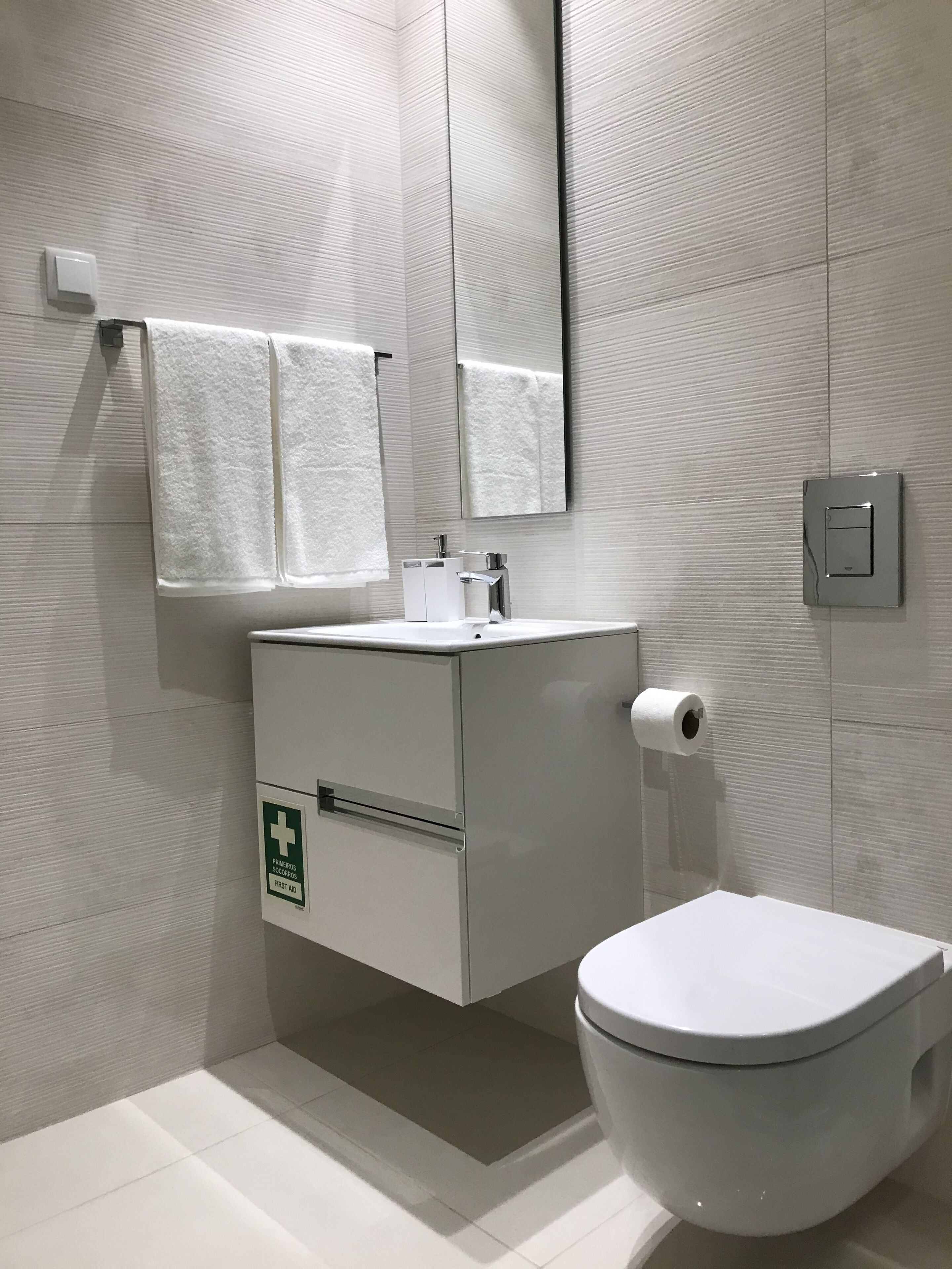 A modern bathroom with a white sink, mirror, and toilet. There is a first aid sign on the cabinet.