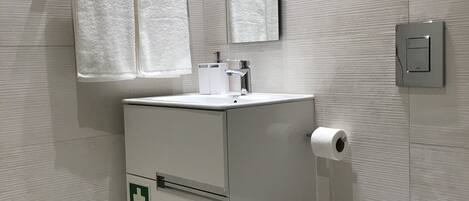 A modern bathroom with a white sink, mirror, and toilet. There is a first aid sign on the cabinet.