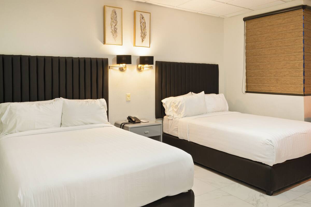 A hotel room with two beds, a nightstand, a telephone, wall-mounted lights, and a window with a blind.