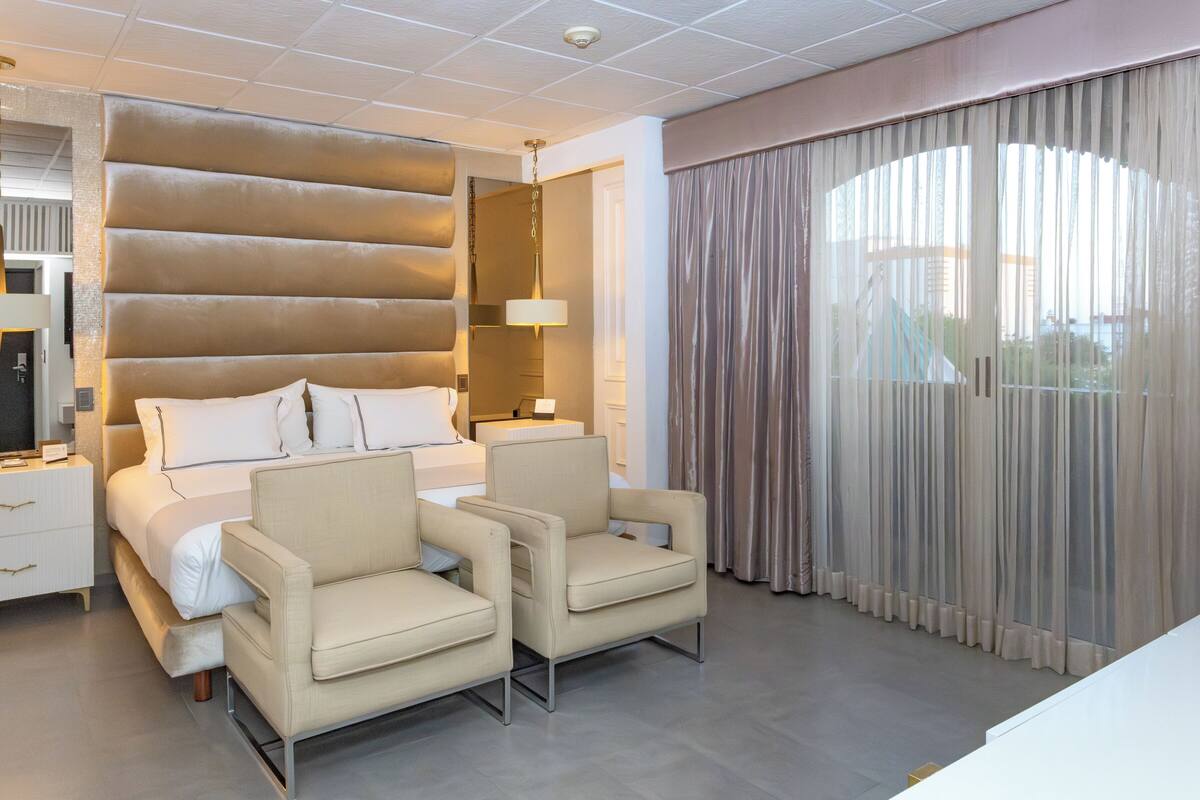 A modern hotel room with a large bed, two armchairs, a bedside table, and a large window with curtains.