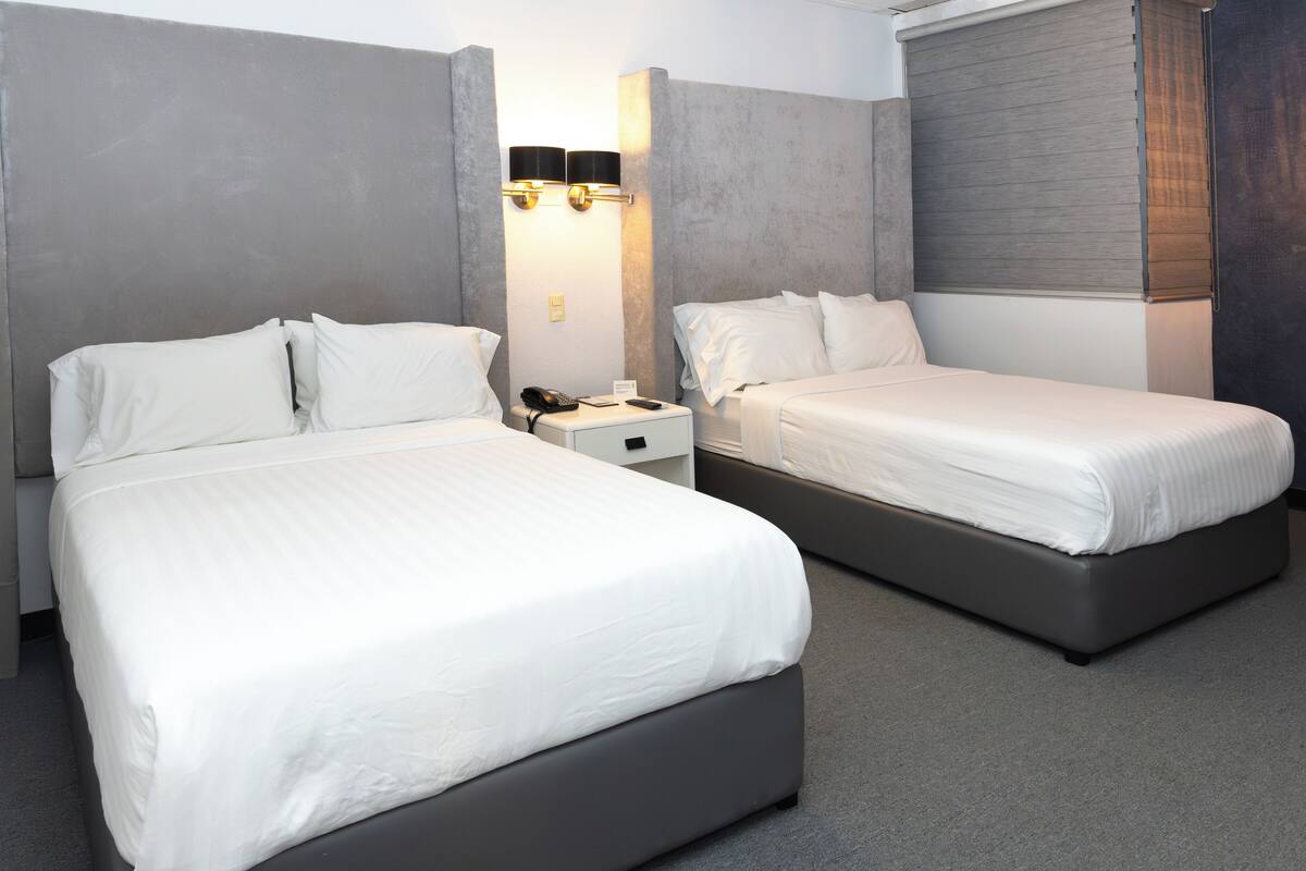 A hotel room with two beds, a nightstand, a lamp, and a window with blinds.