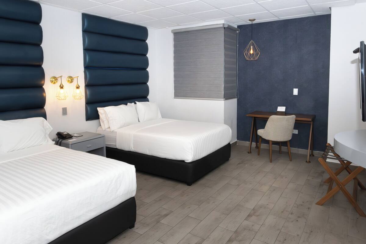A hotel room with two beds, a desk, a chair, a TV, and a window with blinds.