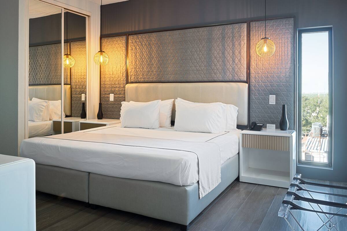 A modern bedroom with a large bed, a bedside table, a mirror, and a view of the city.