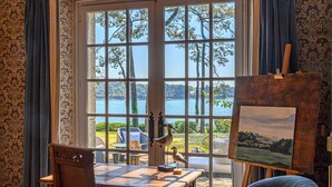 A room with a large window overlooking a lake, a wooden desk, an easel with a painting, and a chair.