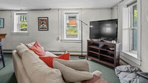 Olohuoneessa on beige sohva, punaiset koristetyynyt, puinen tv-taso ja lattiavalaisin.