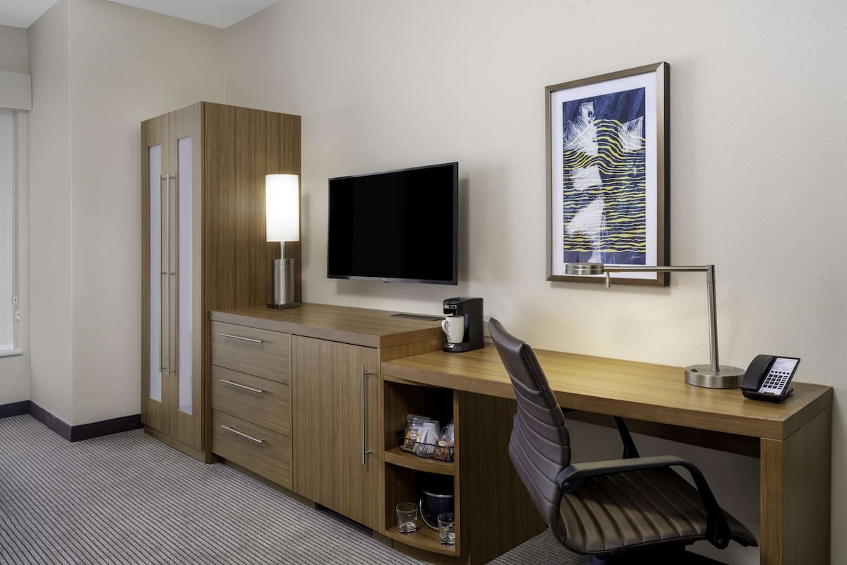 Una habitación de hotel con un escritorio de madera, un televisor de pantalla plana, cafetera, teléfono y una obra de arte enmarcada en la pared.