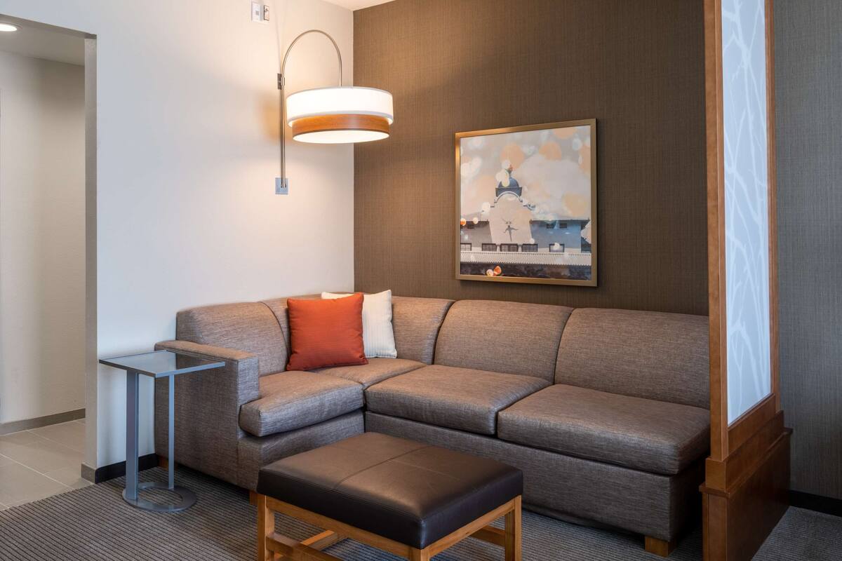 Una habitación de hotel moderna con un sofá modular, una mesita auxiliar pequeña, un banco de madera, una lámpara de pared y un cuadro con una vista urbana enmarcada.