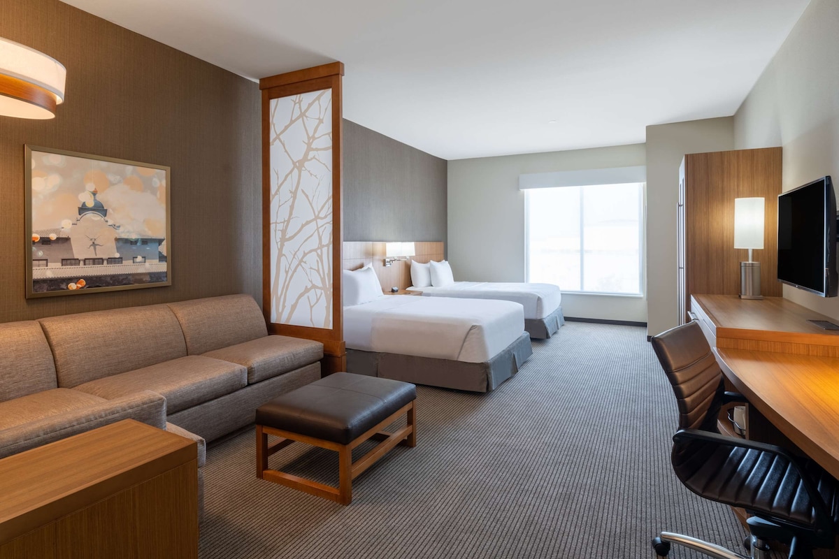 Habitación de hotel con sofá, dos camas individuales, un escritorio con silla, televisor y una ventana grande.