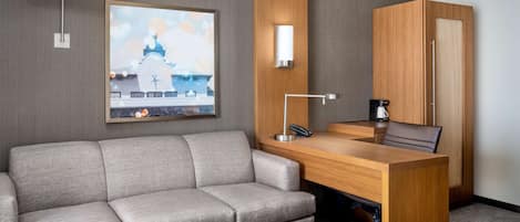 A hotel room with a sofa, a desk with a lamp, a painting of a building, and a wardrobe.