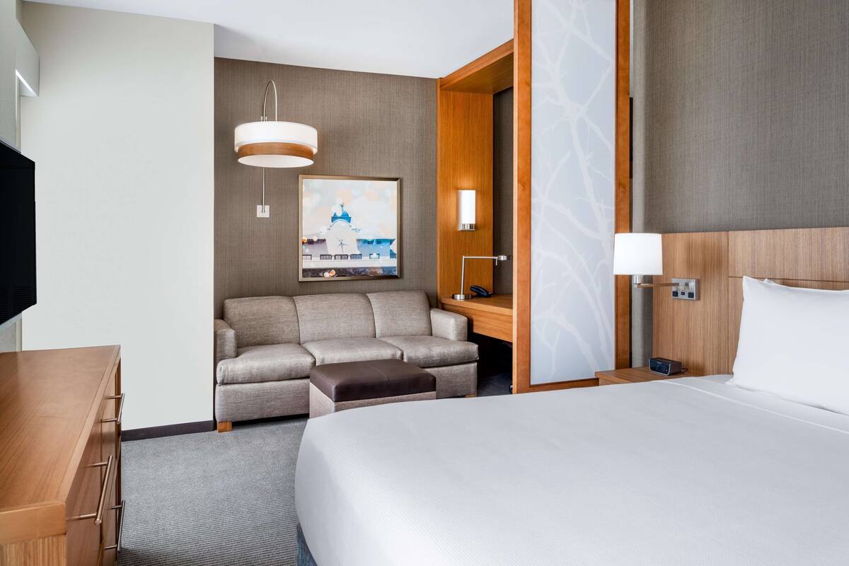 Habitación de hotel con cama, sofá, escritorio, lámpara, cuadro y televisor.
