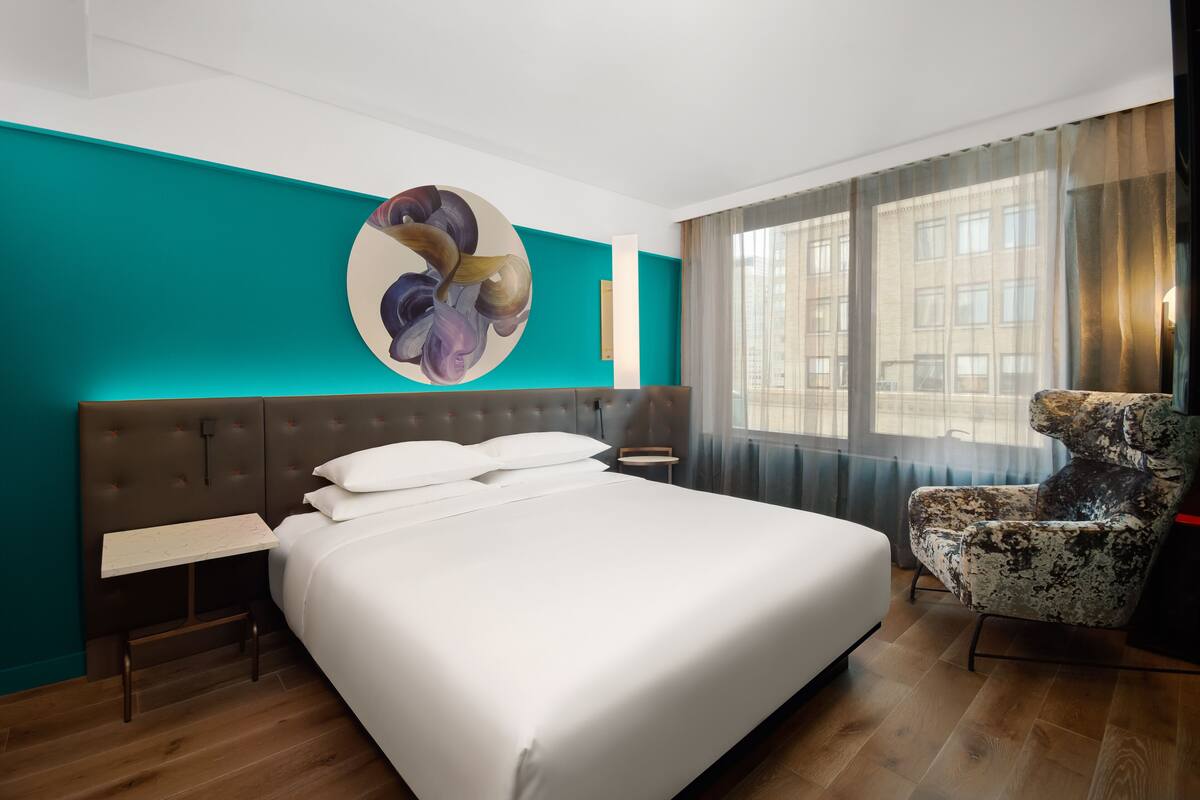 A modern hotel room with a large bed, a side table, a decorative wall piece, and a view of buildings through the window.