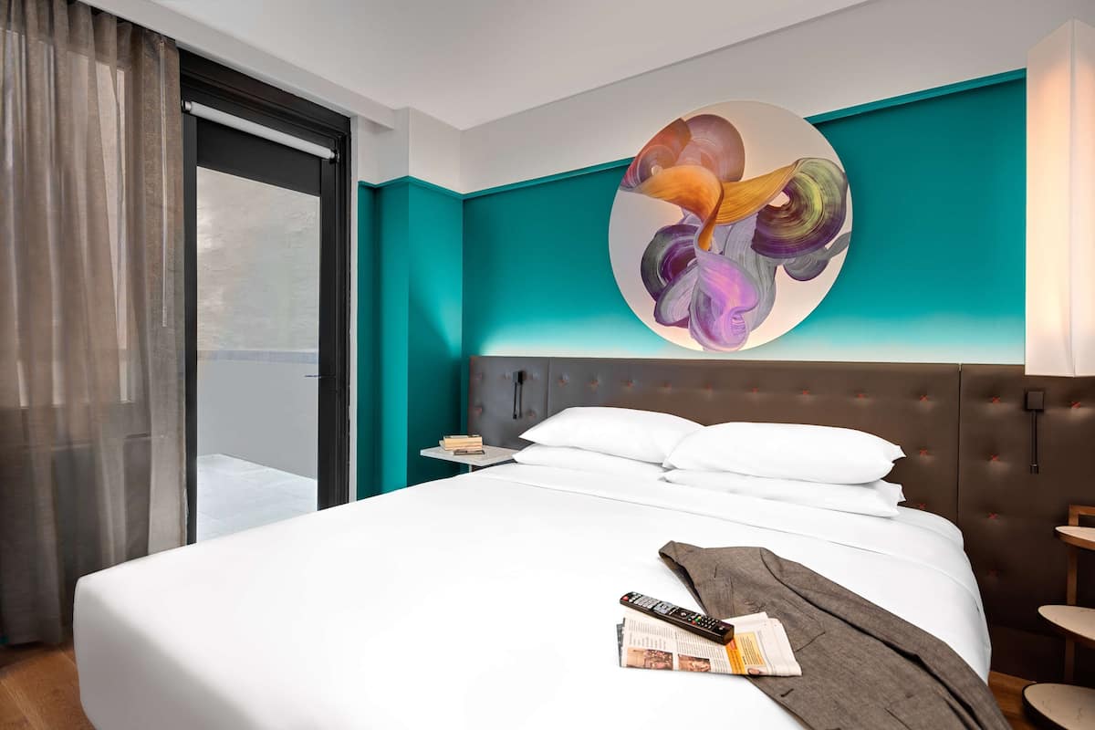 A hotel room with a large bed, a nightstand, a wall art piece, and a view through a window with sheer curtains.