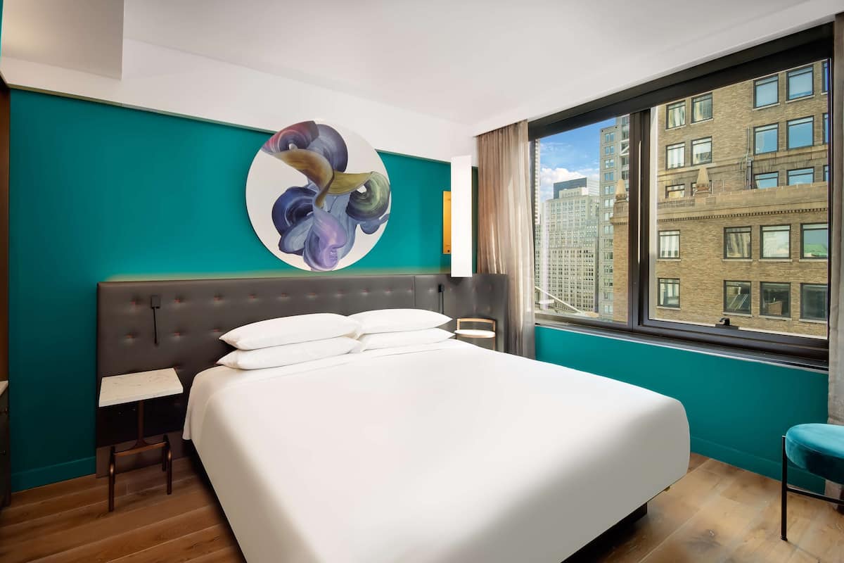 A modern hotel room with a large bed, a small table, a chair, and a city view through a large window.
