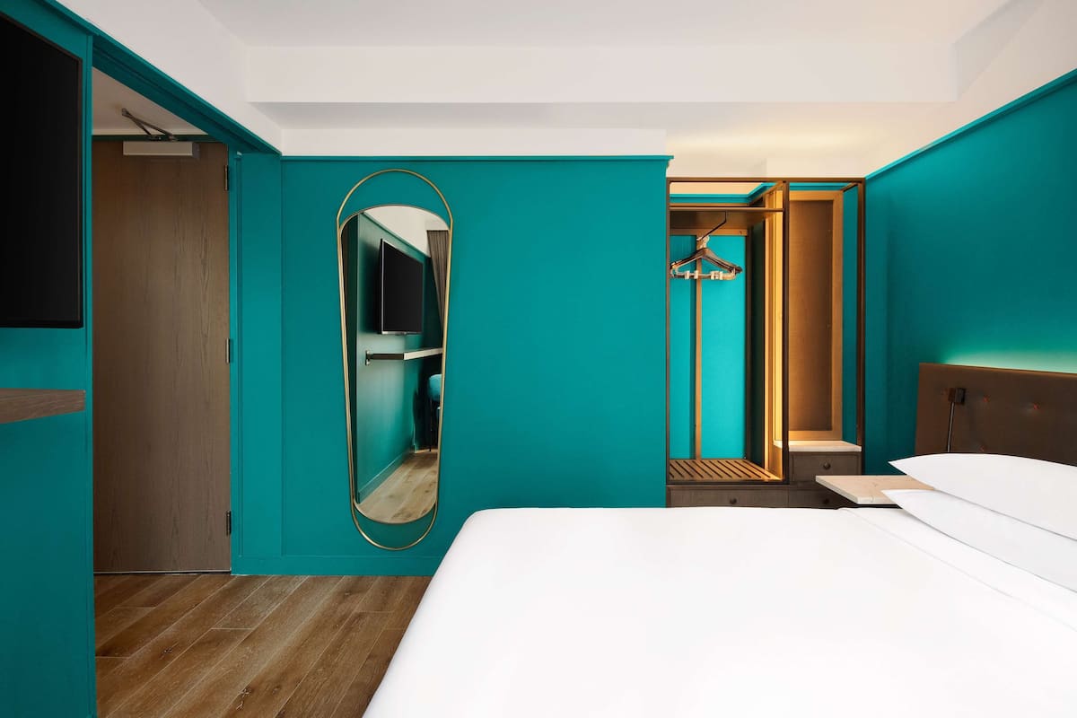 A modern hotel room with teal walls, a large bed, a mirror, a wooden floor, and a walk-in closet.