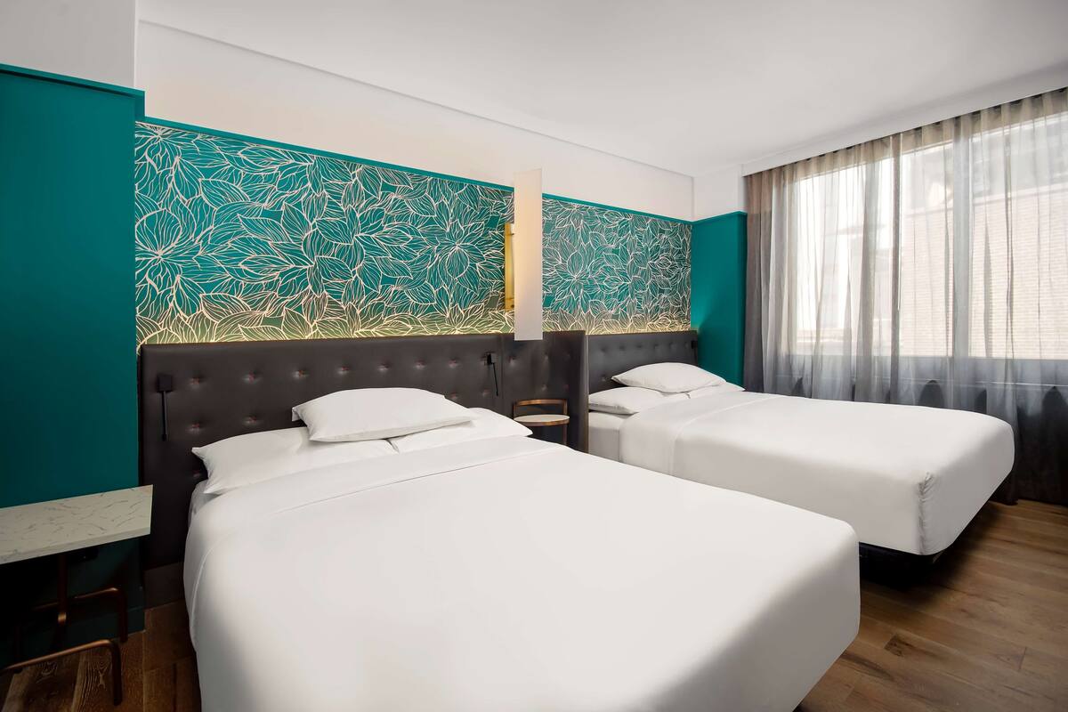 A hotel room with two beds, a teal accent wall, a window with sheer curtains, and a wooden floor.