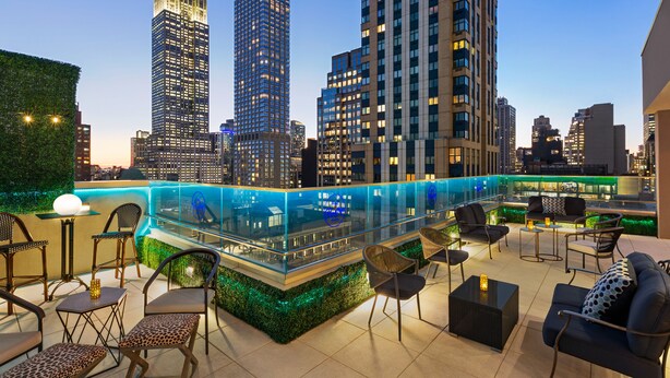 Un bar situé sur le toit, doté d’une rambarde en verre, d’un espace salon avec des chaises et des tables, et offrant une vue sur le panorama urbain en arrière-plan.