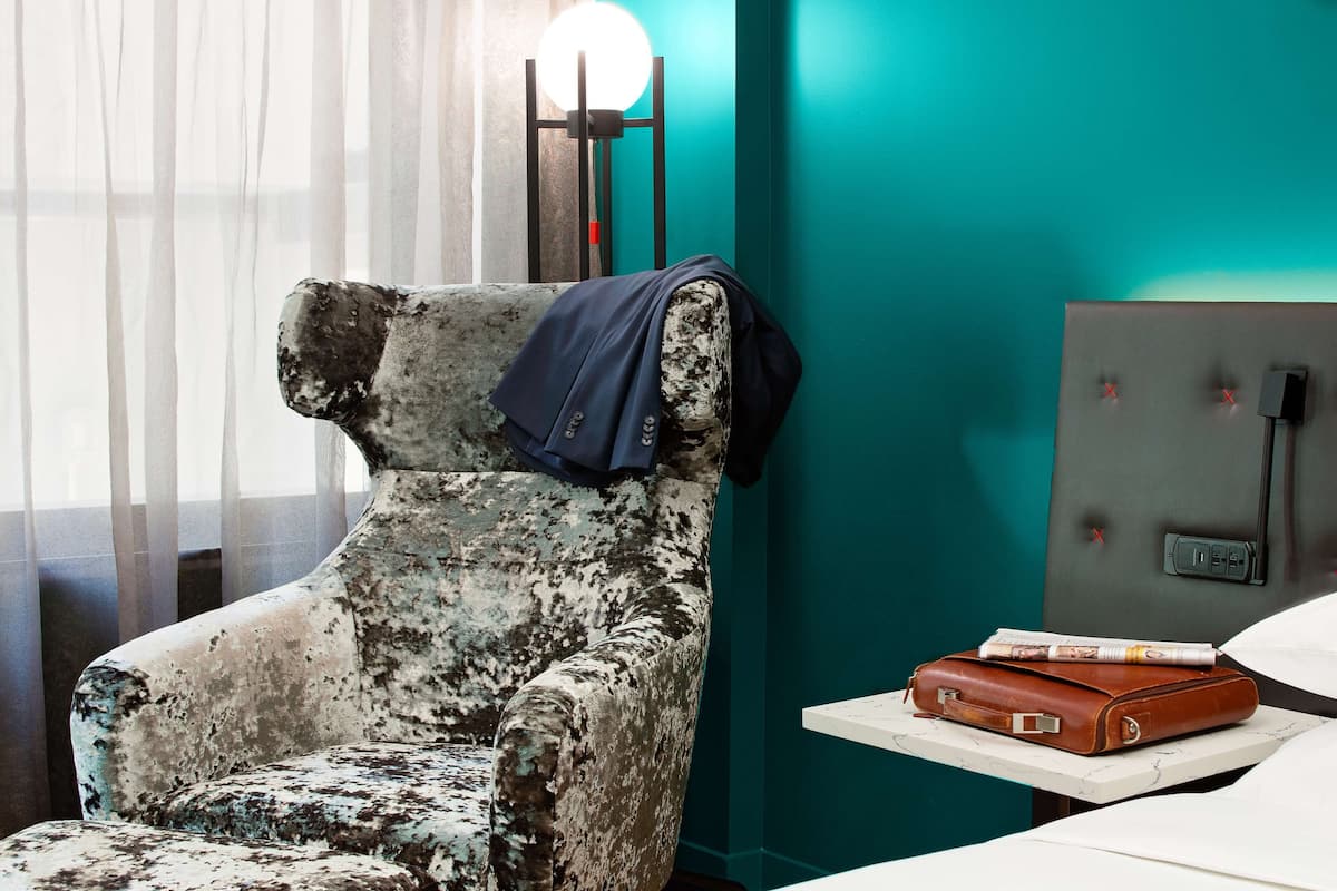 A vintage armchair with a blue throw, a brown leather briefcase, and a magazine on a small table in a room with teal walls and a window with sheer curtains.