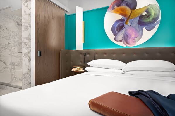 A modern hotel room with a large bed, a wooden headboard, a teal accent wall, and a marble shower area.