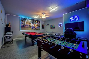 A room with a foosball table, pool table, ceiling fan, and neon gaming sign.