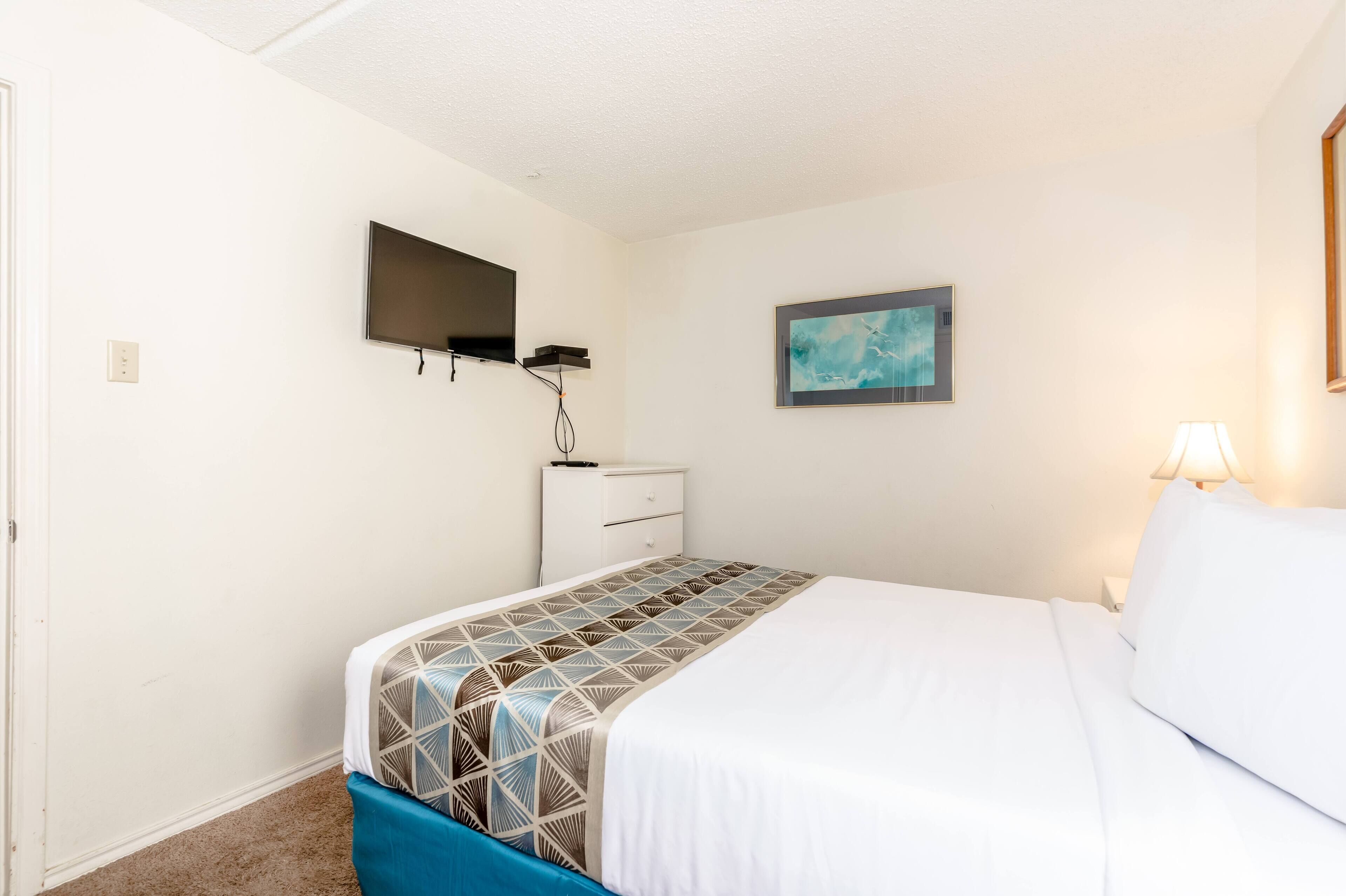 A bedroom with a bed, a TV mounted on the wall, a small dresser, and a painting.