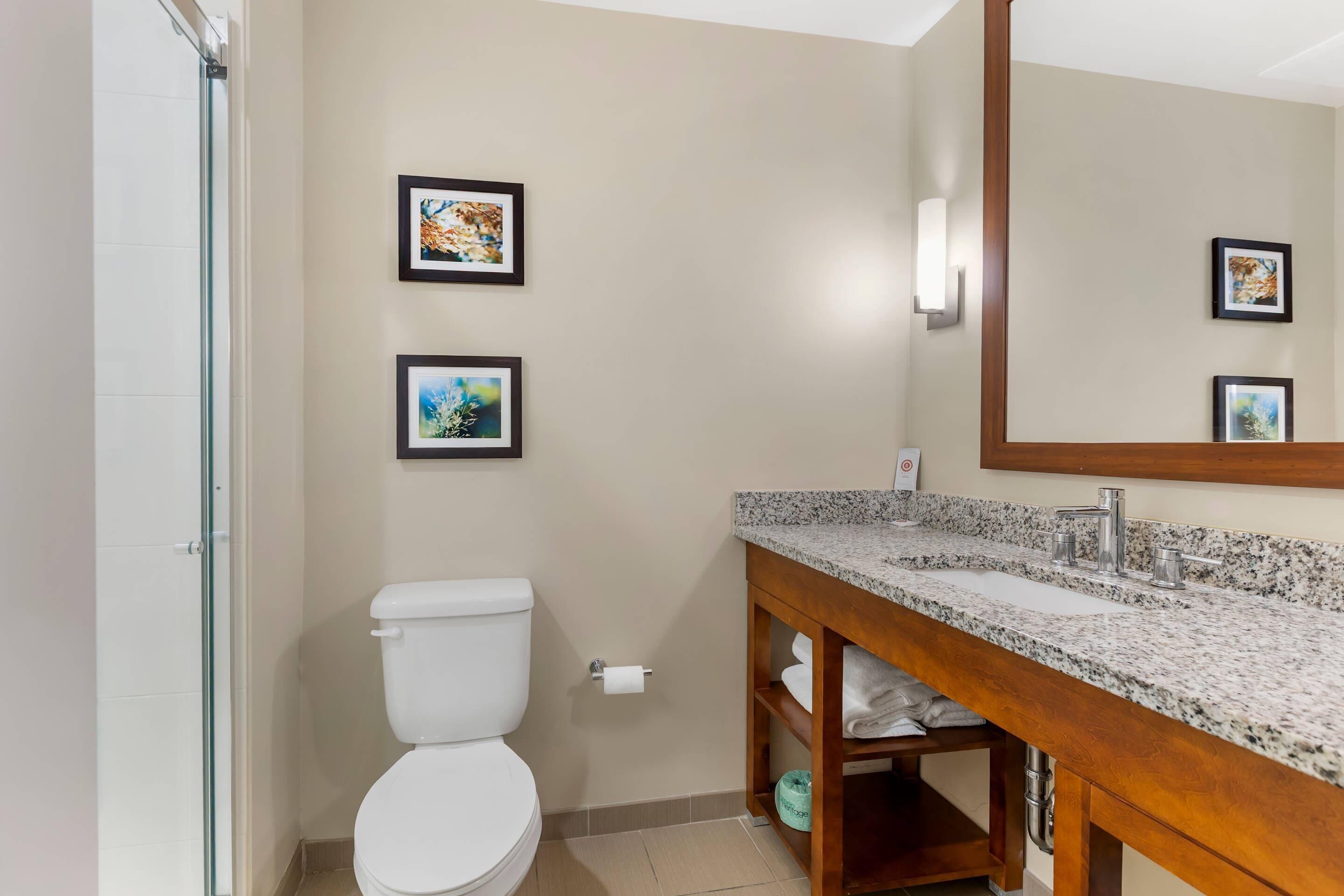 Un baño con inodoro blanco, lavamanos de encimera de granito y mueble de tocador de madera. Hay cuadros enmarcados en la pared.