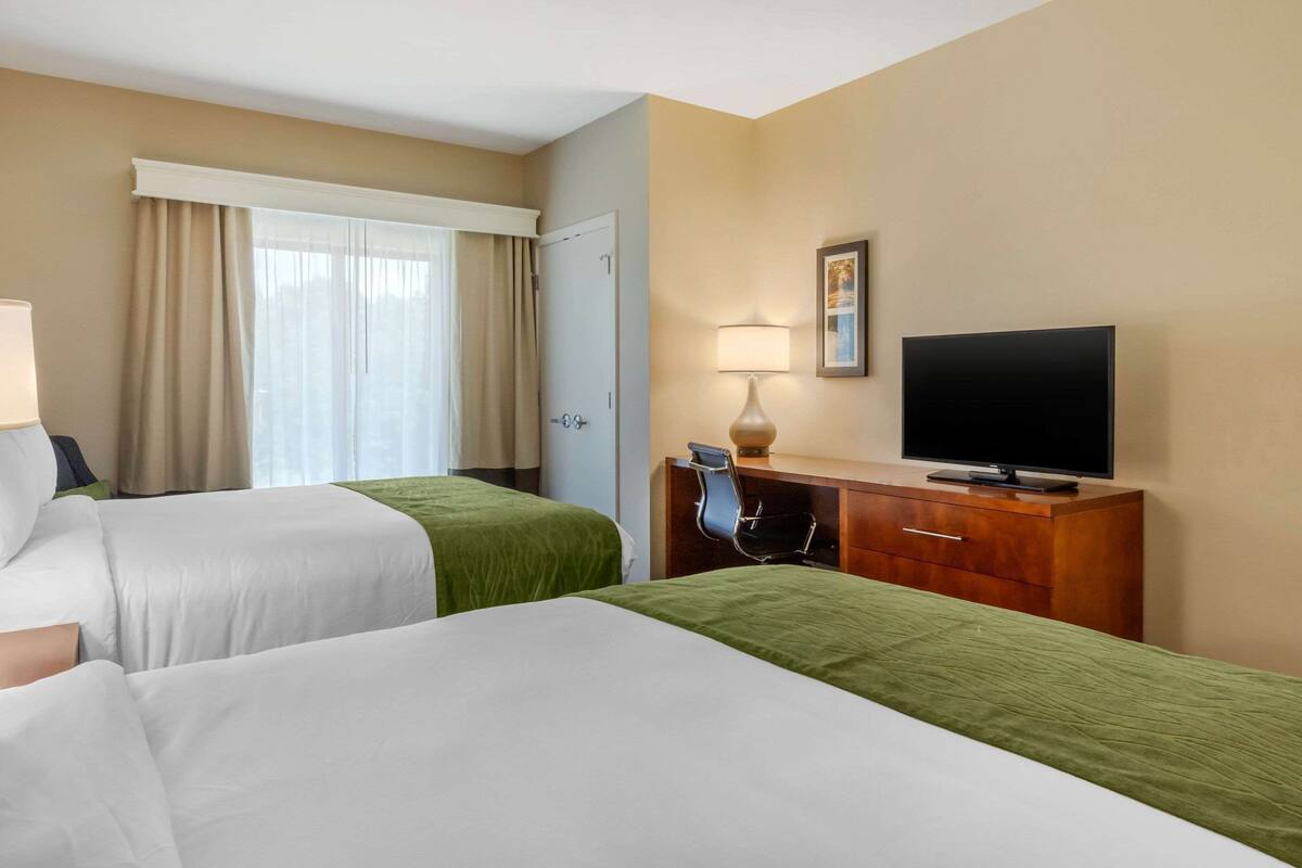 A hotel room with two beds, a television, a desk with a lamp, and a window with curtains.