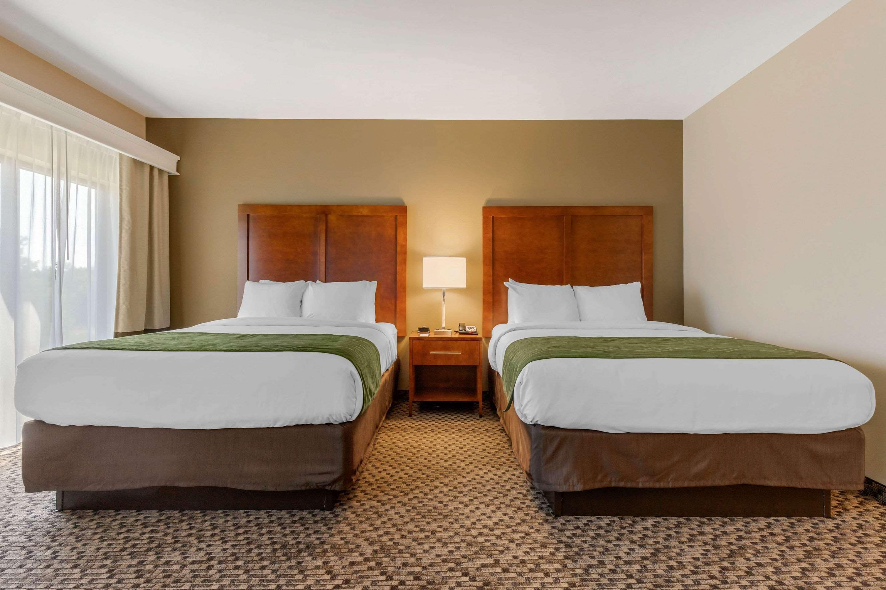 A hotel room with two beds, a nightstand with a lamp, and a window with curtains.