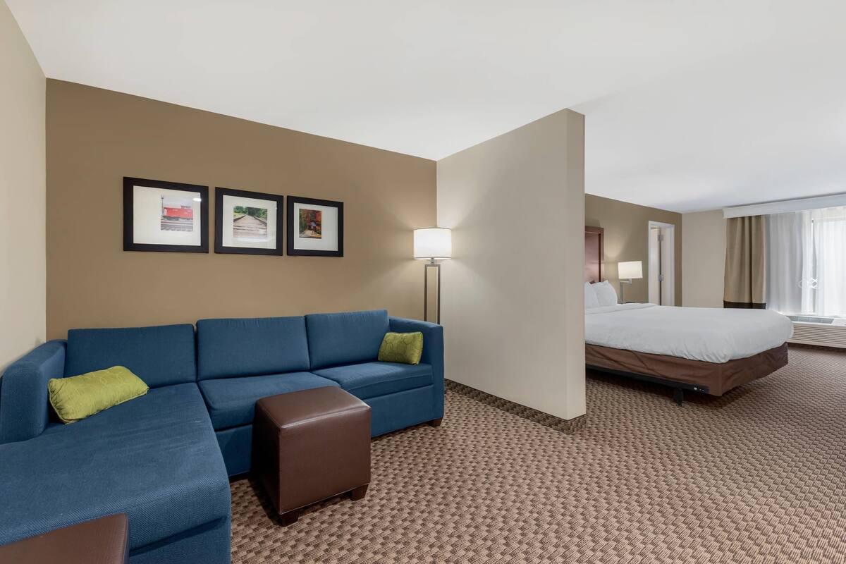 A hotel room with a blue sofa, a brown ottoman, a bed, a lamp, and three framed pictures on the wall.