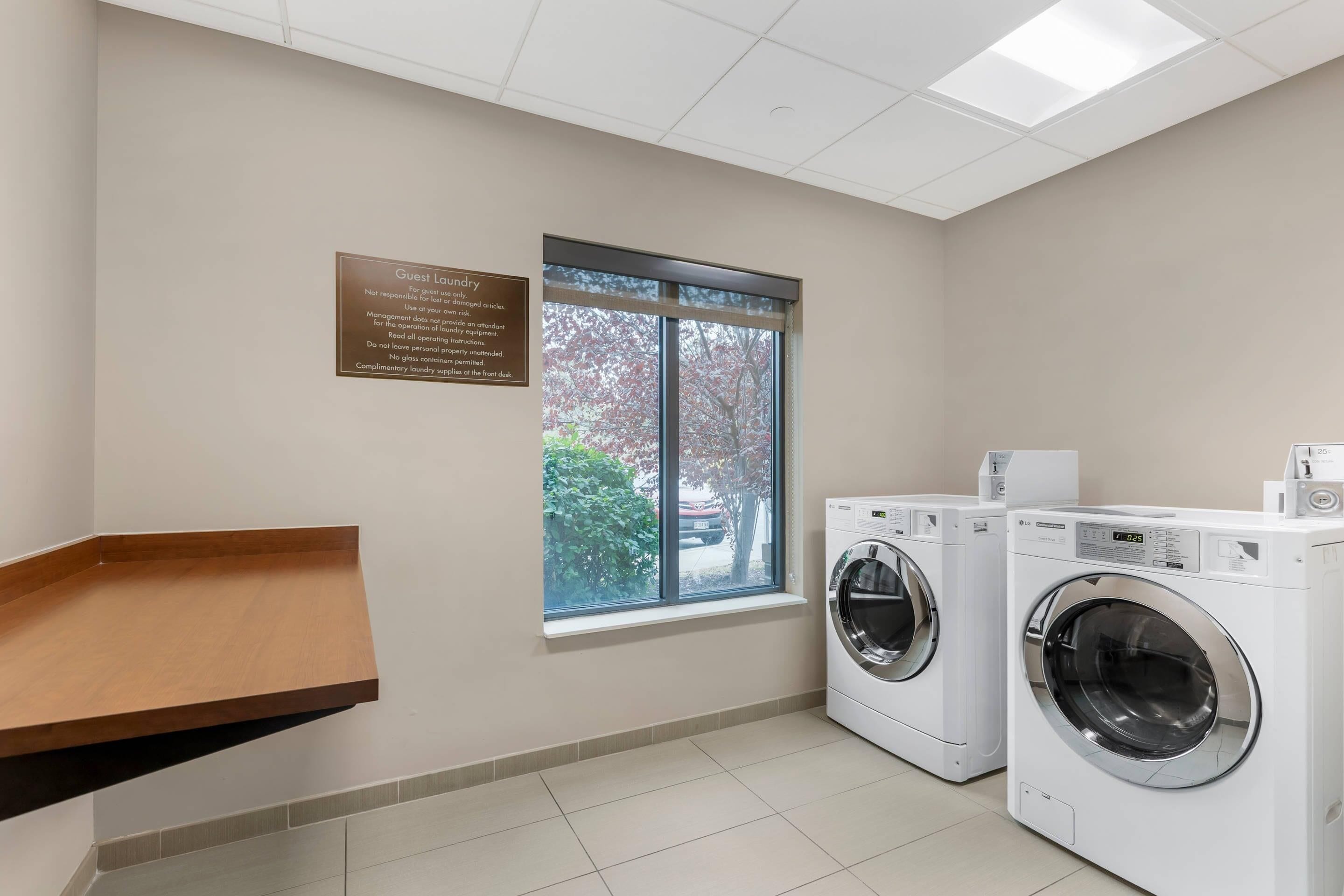A laundry room with two white washing machines, a wooden shelf, a window with a view of trees, and a sign about guest laundry.