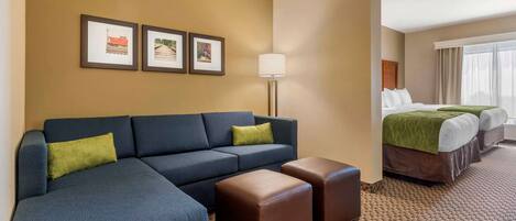 Habitación de hotel con sofá modular, dos otomanas, cama con colcha verde y cuadros en la pared.