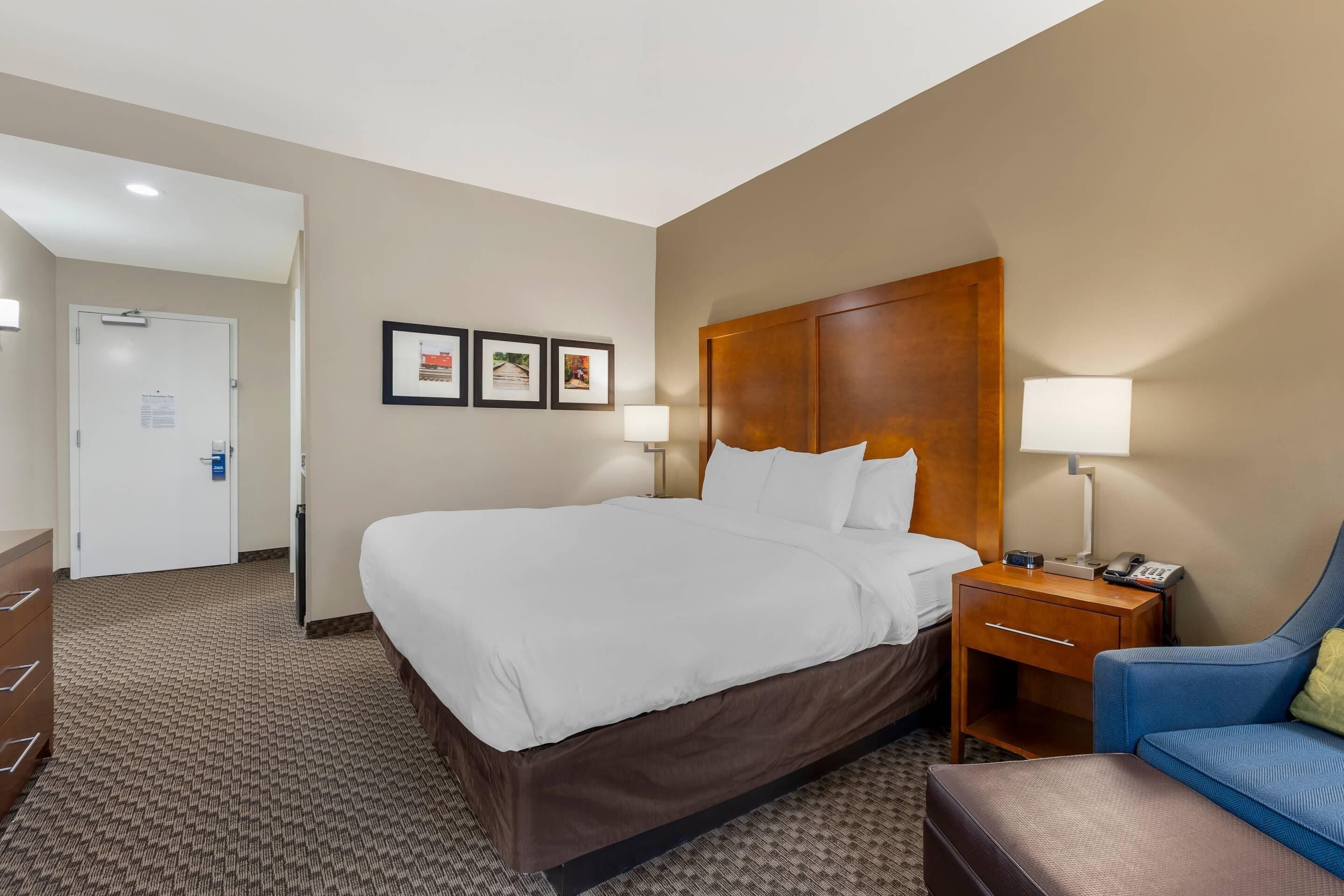 A hotel room with a large bed, a wooden headboard, two bedside tables with lamps, a blue armchair, and a brown sofa.