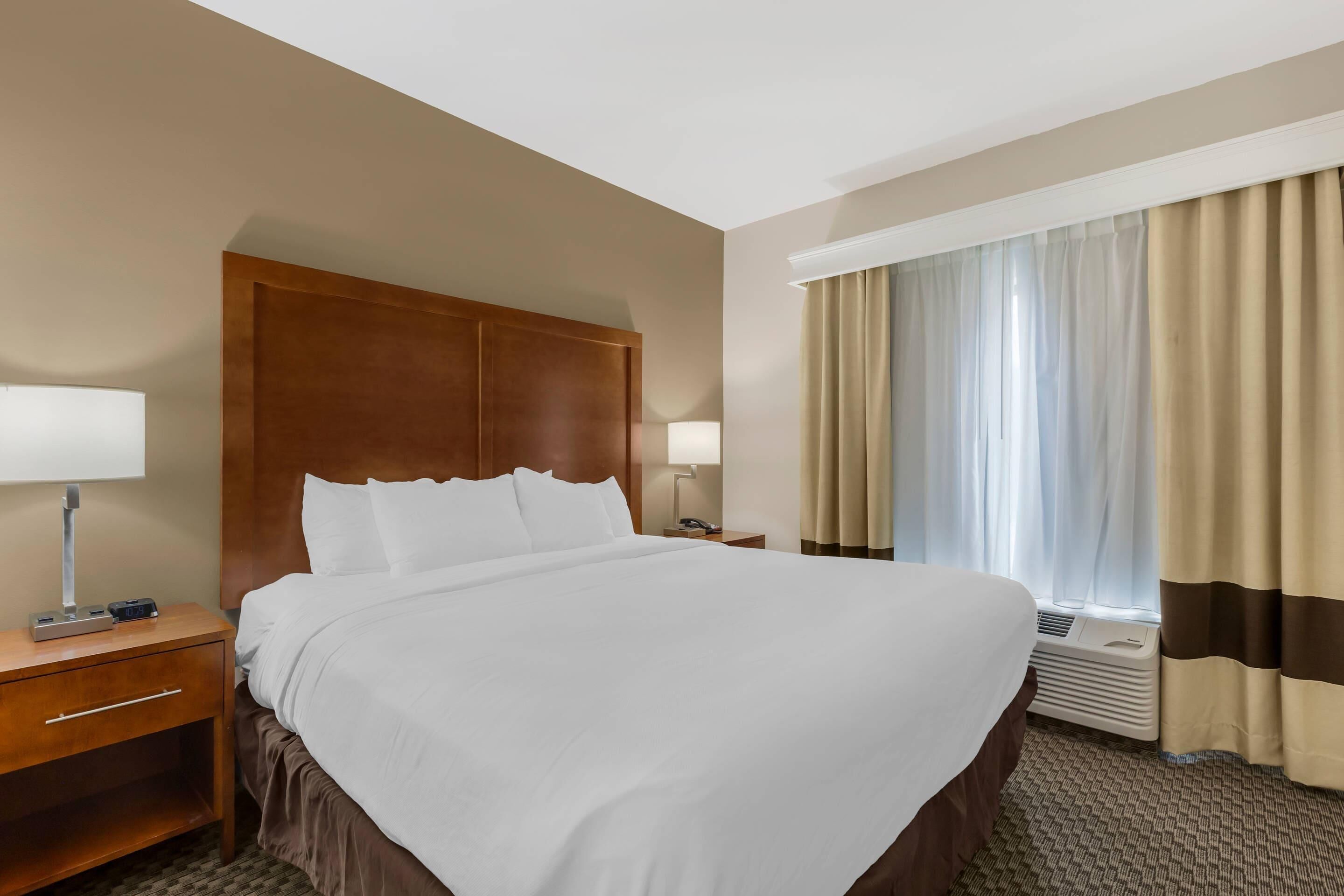 Ein Hotelzimmer mit einem großen Bett, einem hölzernen Kopfteil, einem Nachttisch mit Lampe, einem Fenster mit Vorhängen und einer Klimaanlage.