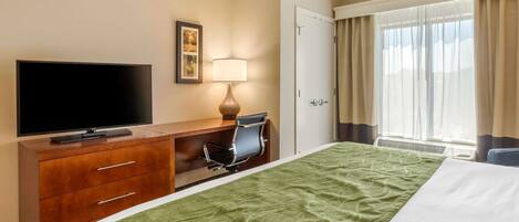 A hotel room with a bed, a desk with a chair, a television, a lamp, and a door.