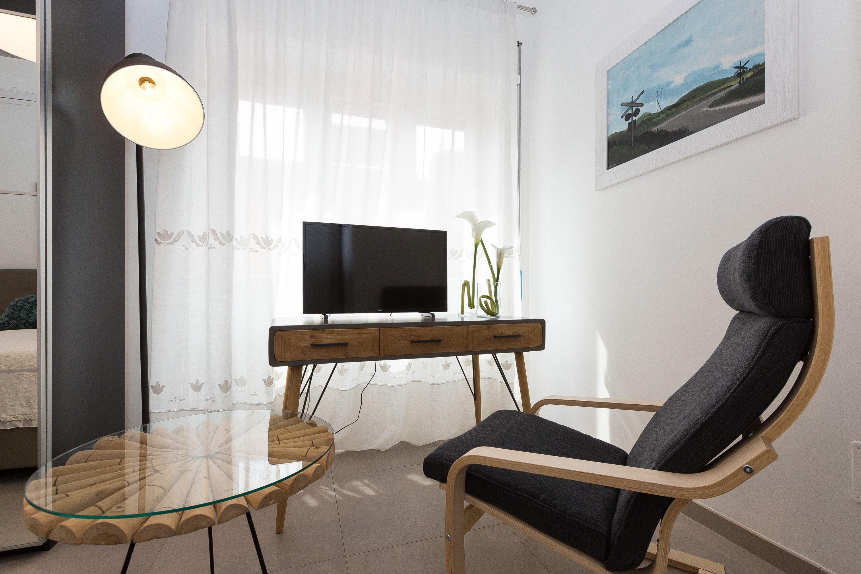 Een moderne woonkamer met een glazen tafel, een zwarte bank, een televisie op een houten kast en een schilderij aan de muur.
