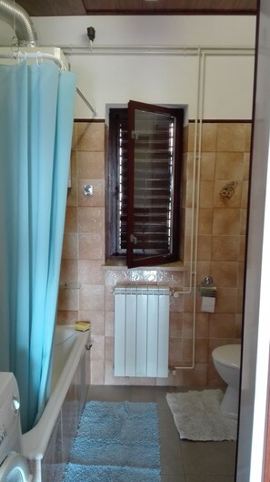 A bathroom with a bathtub, a toilet, a window with blinds, and a radiator.