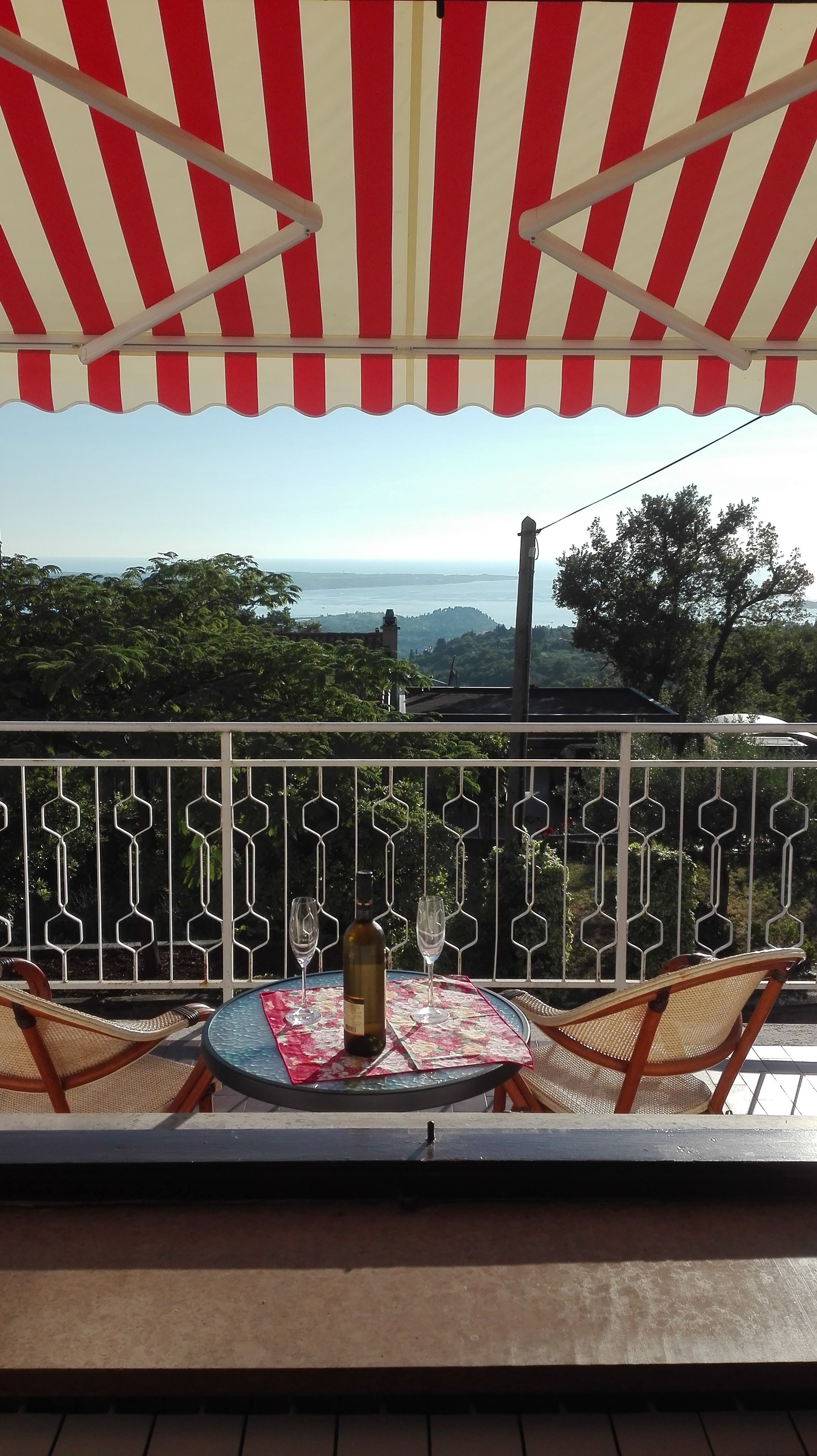 Ein Balkon mit einem rot-weiß gestreiften Sonnensegeltuch, einem Tisch mit einer Flasche und Gläsern sowie Blick auf die Landschaft.