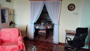 A living room with a red chair, a black office chair, a dining table with a floral tablecloth, and a kitchen area visible through a curtained doorway. - Very Spacious Apartment (Izola)