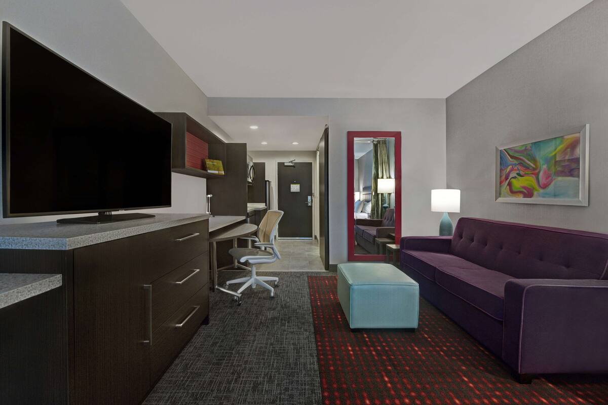 Ein modernes Hotelzimmer mit einem großen Flachbildfernseher, einem Schreibtisch mit Stuhl, einem violetten Sofa und einem kleinen blauen Hocker.