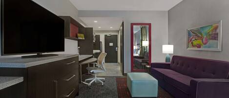 A modern hotel room with a large flat-screen TV, a desk with a chair, a purple sofa, and a small blue ottoman.