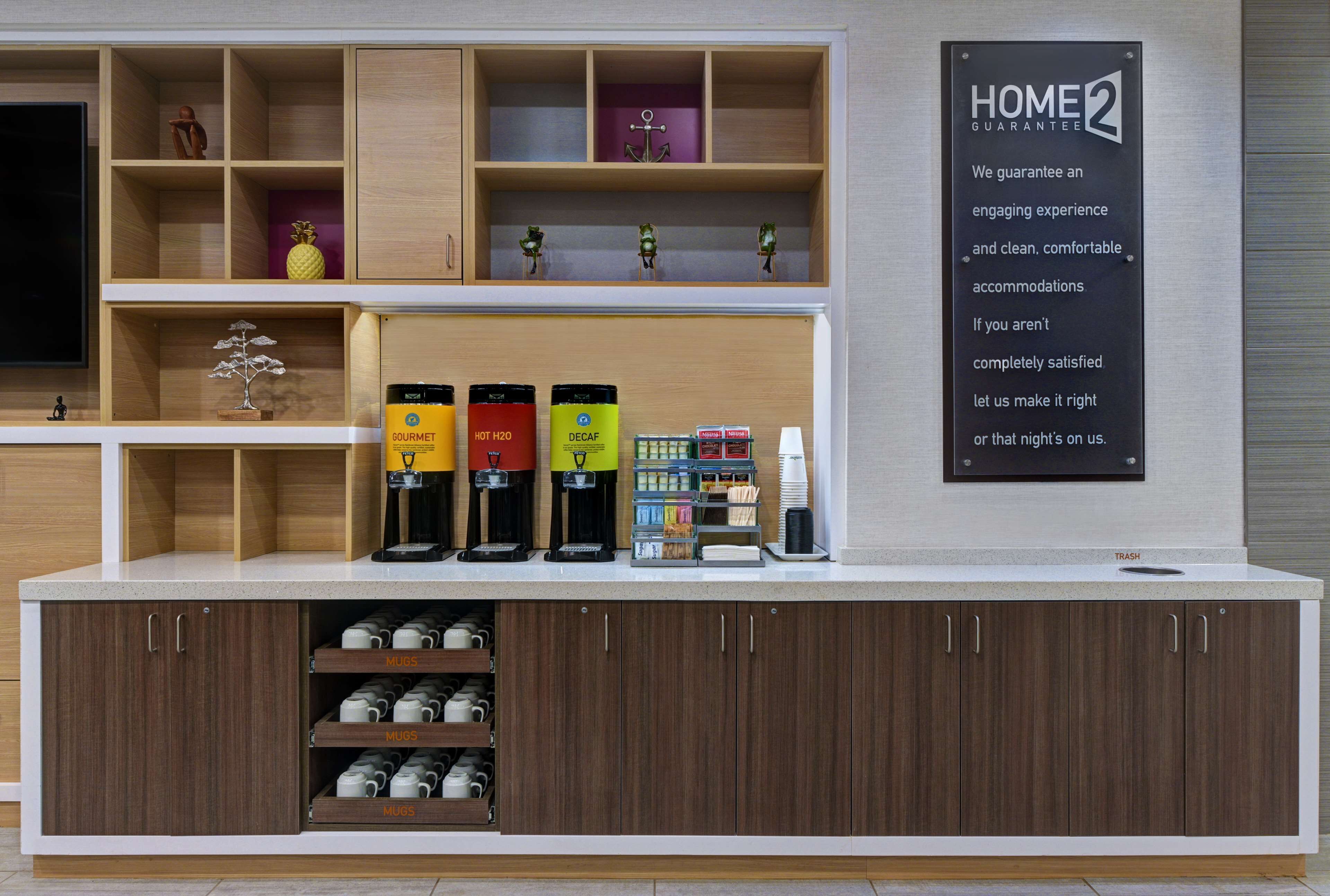 A hotel reception area with a coffee station, mugs, and a sign stating "HOME2 GUARANTEE".
