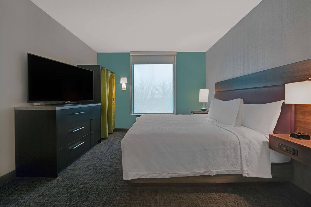 Ein Hotelzimmer mit einem großen Flachbildfernseher, einem Bett mit weißer Bettwäsche, einem Nachttisch mit Lampe und einem Fenster mit Vorhängen.