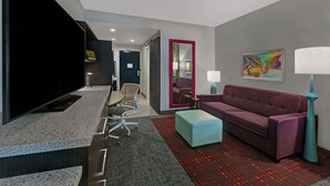 Una habitación de hotel moderna con una televisión de pantalla plana, un sofá púrpura, un reposapiés verde azulado y una alfombra roja estampada.