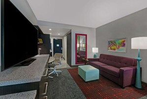 Una habitación de hotel moderna con una televisión de pantalla plana, un sofá púrpura, un reposapiés verde azulado y una alfombra roja estampada.