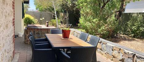 An outdoor patio with a wooden table and wicker chairs, surrounded by a stone wall and greenery.