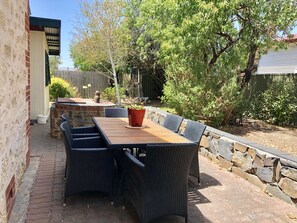 An outdoor patio with a wooden table and wicker chairs, surrounded by a stone wall and greenery.
